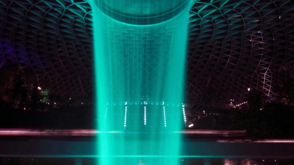 A dark, dramatic shot of the Jewel Changi Rain Vortex at night, illuminated by a brilliant turquoise light shining through the water. This indoor waterfall is a centrepiece of the airport's sensory environment, complete with bespoke audio brand and harmonised soundscapes.